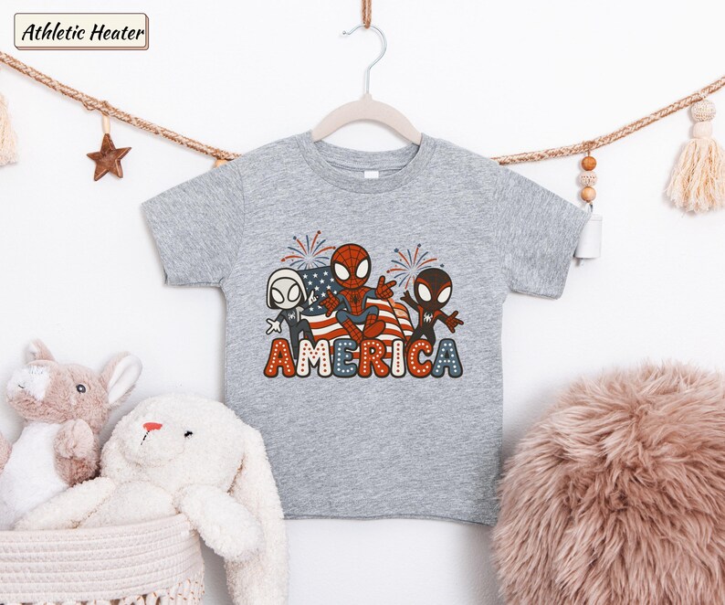 May include: A heather gray short-sleeve t-shirt featuring a graphic of three Spider-Man characters in front of an American flag with fireworks. The word AMERICA is printed below in large, bold, red, white, and blue polka-dotted lettering.