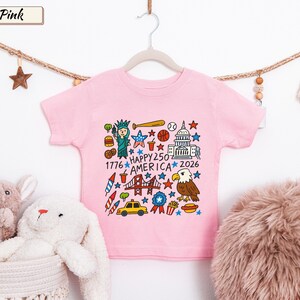 May include: A light pink short-sleeve t-shirt featuring a colorful graphic print of American icons including the Statue of Liberty, a bald eagle, the Capitol building, a taxi, and fireworks with the text Happy 250 America 1776 2026.