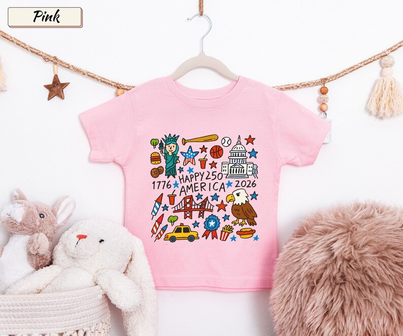 May include: A light pink short-sleeve t-shirt featuring a colorful graphic print of American icons including the Statue of Liberty, a bald eagle, the Capitol building, a taxi, and fireworks with the text Happy 250 America 1776 2026.