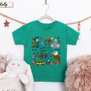 May include: A kelly green short-sleeve t-shirt featuring a colorful graphic print of American icons including the Statue of Liberty, a bald eagle, the Capitol building, a taxi, and fireworks, with the text "HAPPY 250 AMERICA 1776 2026".