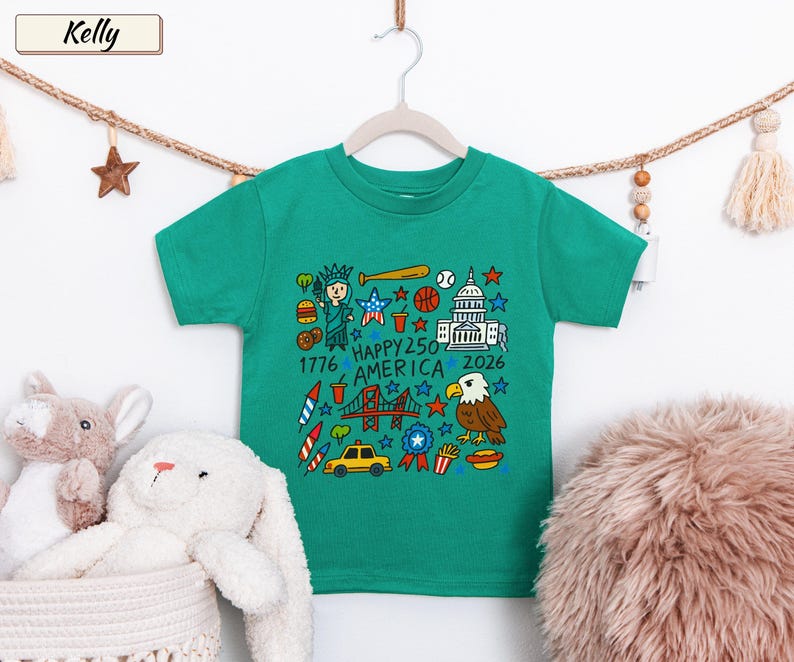 May include: A kelly green short-sleeve t-shirt featuring a colorful graphic print of American icons including the Statue of Liberty, a bald eagle, the Capitol building, a taxi, and fireworks, with the text "HAPPY 250 AMERICA 1776 2026".