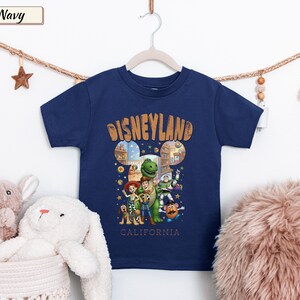 May include: Navy blue t-shirt featuring a colorful graphic of Disney characters with the text "Disneyland California". The design includes images of Woody, Buzz Lightyear, and other characters. The shirt is hanging on a white hanger.