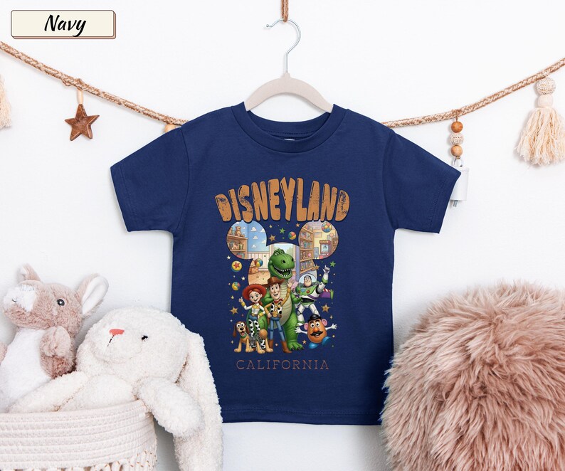 May include: Navy blue t-shirt featuring a colorful graphic of Disney characters with the text "Disneyland California". The design includes images of Woody, Buzz Lightyear, and other characters. The shirt is hanging on a white hanger.
