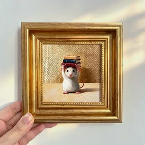Può includere: Un piccolo dipinto incorniciato raffigurante un topo bianco in piedi sulle zampe posteriori che bilancia una pila di tre libri sulla testa. Lo sfondo è una superficie dorata testurizzata e la cornice è in materiale decorativo color oro.