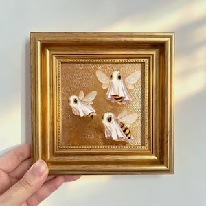 May include: A square, ornate gold-colored frame containing a painting of three bees, each draped in a white sheet resembling a ghost costume. The background of the painting is a textured, metallic gold surface with a beaded inner border detail.