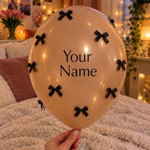 May include: A beige balloon featuring the black text Your Name printed in the center, decorated with several small black ribbon bows attached to the surface of the balloon in a scattered pattern across the rounded exterior.