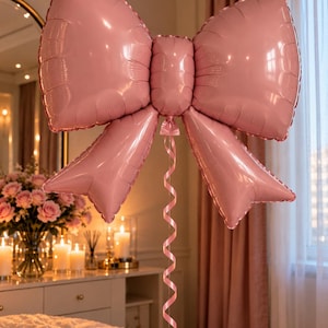 May include: A large, glossy, light pink foil balloon shaped like a decorative bow. The balloon features a central knot with two wide loops and two trailing ribbon ends. A thin, matching pink curling ribbon hangs down from the center knot.
