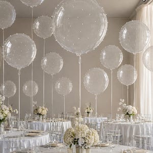 May include: A collection of clear, spherical balloons decorated with small white pearls, each attached to a long white ribbon. These balloons are suspended above round dining tables set with white tablecloths and white floral centerpieces.