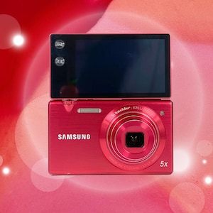 May include: A bright red Samsung digital camera with a 5x zoom lens. The camera has a large LCD screen and a Schneider-Kreuznach lens. The camera is on a red and pink background.