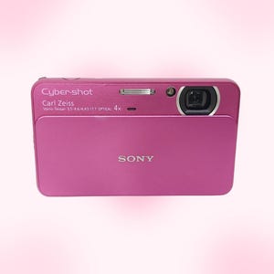 Puede incluir: Cámara digital Sony Cyber-shot magenta. La cámara tiene una lente Carl Zeiss y presenta el texto "Cyber-shot" y "SONY". La cámara tiene un flash y una lente en la parte delantera.