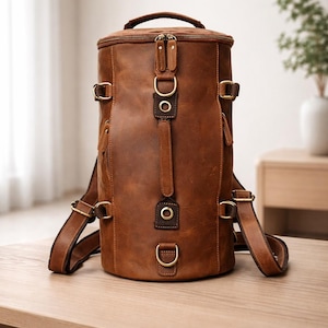 Può includere: Uno zaino cilindrico in pelle marrone con spallacci regolabili e una maniglia superiore. La borsa presenta fibbie e anelli in metallo color ottone. Lo zaino è appoggiato su un tavolo di legno color chiaro.