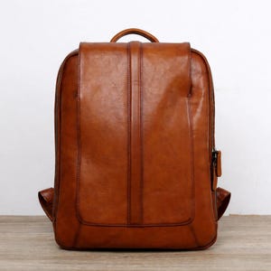 May include: A brown leather backpack with a rectangular shape and a flap closure. The backpack has a top handle and two adjustable shoulder straps. The leather has a distressed look and a zipper closure on the side.