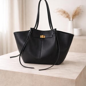 May include: Black leather tote bag with a gold-tone clasp. The bag has two top handles and long, thin straps that tie at the front. The bag is sitting on a light-colored surface.