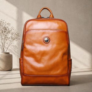 May include: A brown leather backpack with a top handle and a front pocket. The backpack has a circular emblem on the front and two side pockets. The leather has a glossy finish.