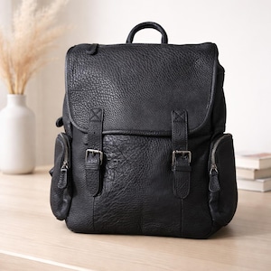 Könnte beinhalten: Schwarzer Lederrucksack mit strukturierter Oberfläche. Mit Klappenverschluss mit zwei Schnallenriemen, zwei seitlichen Reißverschlusstaschen und einem oberen Griff. Die Tasche steht auf einer Holzoberfläche.