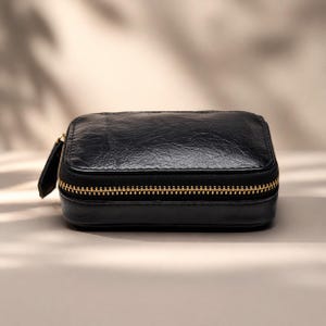 May include: A small, rectangular black leather case with a gold zipper closure. The case has a textured surface and a small, attached zipper pull. The overall design is simple and elegant, suitable for storing small items.