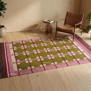 May include: A rectangular area rug featuring a repeating grid pattern in olive green, cream, and pink. The rug has a border of pink and cream stripes, with dark brown fringe detailing along the short edges. The design includes small white rectangular accents.