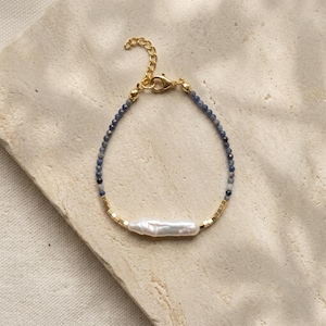 May include: A delicate bracelet featuring a central elongated white baroque pearl flanked by small gold-toned square beads. The remainder of the strand consists of tiny faceted blue-grey gemstone beads, finished with a gold-toned lobster clasp.