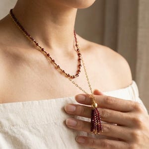 Op de afbeelding: Een gelaagde ketting met een fijne goudkleurige ketting en een streng van kleine, gefacetteerde donkerrode granaatkralen. De ketting heeft een centraal goudkleurig accent waaraan een kwastje van bijpassende rode granaatkralen hangt.