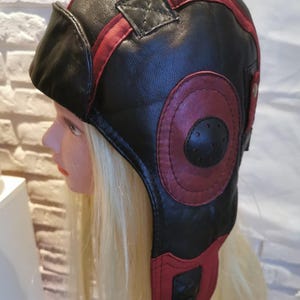 Può includere: Cappello da aviatore in pelle nera con dettagli rossi. Il cappello presenta una copertura auricolare arrotondata, una visiera anteriore e un cerchio rosso decorativo sul lato. Una striscia rossa corre lungo la parte superiore. Il cappello è fissato con una fibbia.