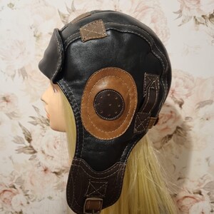 Op de afbeelding: Zwarte en bruine leren pilotenmuts met oorflappen en een kinband. De hoed heeft bruine cirkelvormige patches aan de zijkanten en decoratieve stiksels. De hoed is ontworpen met een vintage esthetiek.
