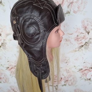 Può includere: Un cappello da aviatore marrone scuro caratterizzato da una costruzione a più pannelli con cuciture chiare visibili. Il cappello include paraorecchie laterali, un dettaglio circolare applicato sul lato e un piccolo accento metallico.