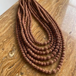 Puede incluir: Una colección de collares de cuentas marrones, probablemente cuentas de mala, dispuestas en un patrón en capas. Las cuentas son de un tono cálido y terroso, y los collares están conectados en la parte superior con un cierre dorado. El fondo es una superficie clara y texturizada.