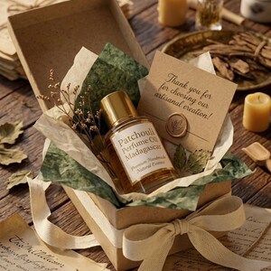 Puede incluir: Un frasco de perfume "Patchouli Perfume Oil Madagascar" se encuentra en una caja de regalo abierta con una cinta crema. La caja contiene papel verde y crema, flores secas y una tarjeta con el texto "¡Gracias por elegir nuestra creación artesanal!"