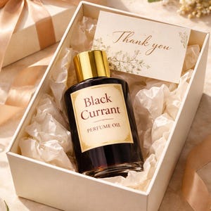 Puede incluir: Un frasco de aceite de perfume "Black Currant" con tapa dorada se encuentra en una caja de regalo blanca con papel de seda. También se ve una tarjeta de "Gracias" y una caja de regalo envuelta con una cinta dorada.