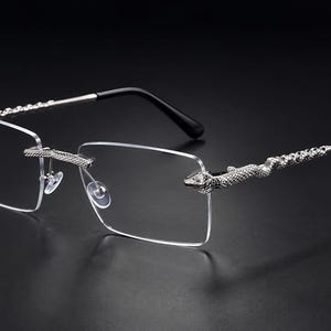 May include: Rimless rectangular eyeglasses featuring silver-toned metal temples shaped like textured snakes. The bridge and hinges also display intricate snake-inspired detailing. The temple tips are finished with smooth black plastic covers.