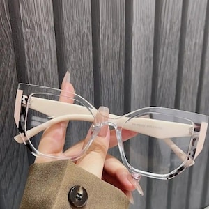 May include: Clear eyeglasses with a cream-colored frame and a subtle leopard print accent on the outer corners. The glasses have a modern, angular shape and are held by a person.