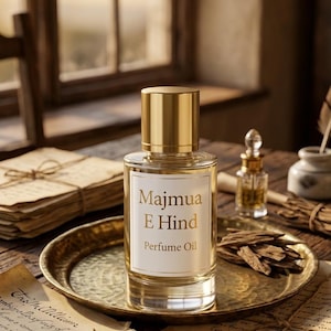 May include: A clear glass perfume oil bottle with a gold cap and label that reads "Majmua E Hind Perfume Oil" sits on a gold tray. The image includes a small glass bottle, wood pieces, and aged paper, suggesting a vintage aesthetic.