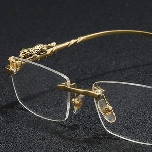 May include: Rimless spectacles with gold-coloured frames. The temples feature a twisted design and a decorative leopard figure near the hinges. The lenses are clear, and the nose bridge has adjustable pads. The spectacles are set against a dark background.