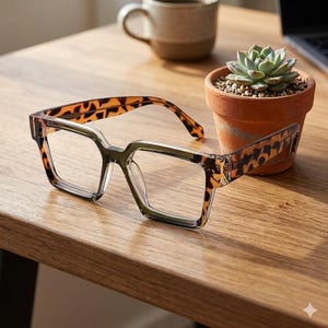 May include: Eyeglasses with a square frame and a tortoiseshell pattern on the arms. The clear frame glasses are on a wooden surface, with a small potted succulent and a coffee mug in the background.
