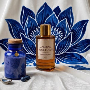 Puede incluir: Composición con un tarro de cristal azul, un frasco de aceite de perfume etiquetado "Calming Blue Lotus" y arándanos. El fondo es una tela blanca con un gran diseño de flor de loto azul.