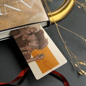 Può includere: Una copia di "The Vampire Armand" di Anne Rice con un segnalibro con un'illustrazione di due persone. Il libro è legato con un nastro rosso. Sono presenti anche occhiali da sole rotondi e un quadro con cornice dorata.