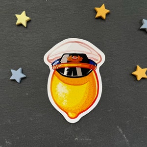 Könnte beinhalten: Eine gelbe Zitrone, die einen weißen und blauen Kapitänshut mit einem roten Emblem trägt. Die Zitrone befindet sich auf einem schwarzen Hintergrund mit gelben Sternen.