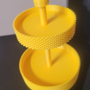 May include: A bright yellow, two-tiered serving stand. The stand has a central post with a mushroom-shaped top. Each tier is a circular tray with a textured, diamond-patterned edge. The stand is made of plastic.