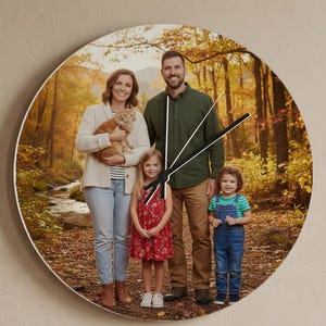 May include: A round wall clock featuring a family portrait set against a backdrop of a forest with autumn foliage. The clock has black hands and a white border. The clock is mounted on a beige wall.