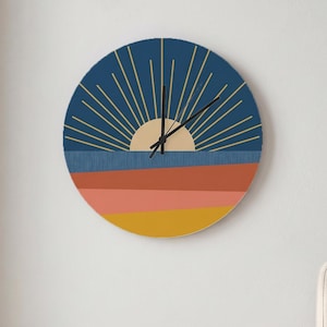 May include: A circular wall clock featuring a graphic design of a sun rising over a landscape. The clock face displays a dark blue sky with radiating gold lines, a beige sun, and horizontal bands of blue, terracotta, pink, and mustard yellow.