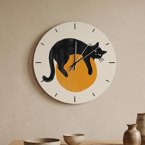 May include: A round wall clock with a cream-colored face featuring a black cat illustration draped over a solid orange circle. The clock has black hour markers and thin black hands. The design is centered on the circular clock face.