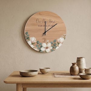 Könnte beinhalten: Runde Wanduhr aus Holz mit floralen Akzenten. Das Zifferblatt zeigt den Text "Our New Home" und "SARAH & MARK". Weiße Blumen und grüne Blätter schmücken den unteren Teil. Schwarze Zeiger zeigen die Uhrzeit an.