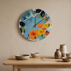 Puede incluir: Un reloj de pared redondo con un diseño floral abstracto y colorido con flores naranjas, amarillas y rojas sobre un fondo azul. La esfera muestra números negros del 1 al 12 e incluye dos manecillas negras para indicar la hora.
