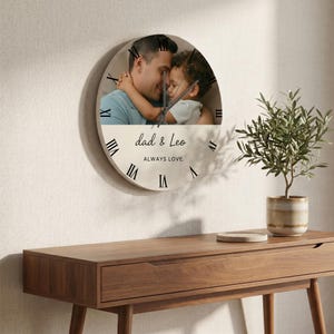 May include: Circular wall clock with a photograph of a father and child. The clock face has black Roman numerals and the text "dad & Leo ALWAYS LOVE." The clock is mounted on a beige wall, with a wooden table and a plant in the foreground.