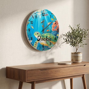 May include: Round wall clock featuring a colorful underwater scene with a sea turtle, fish, and coral. The clock face has black numerals and hands, with the name "Jackie" in script. The clock is mounted on a light-colored wall.