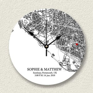 May include: A round white wall clock featuring a black and white map design. The clock face includes the names "SOPHIE & MATTHEW", location details, and a date. A red heart marks a specific location on the map. Black clock hands indicate the time.