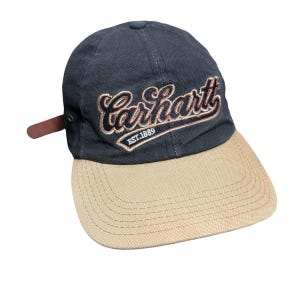 Puede incluir: Una gorra de béisbol azul marino con visera color canela y una correa de cuero marrón en el lateral. El frente presenta el texto bordado Carhartt en fuente cursiva con un contorno canela, y el texto EST.1889 en blanco debajo sobre fondo canela.