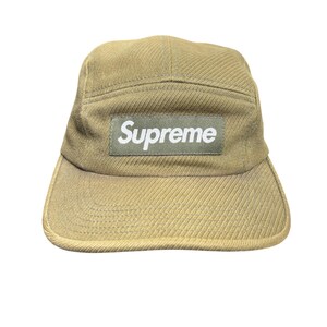 May include: An olive green five-panel cap featuring a rectangular patch on the front. The patch is a darker olive green shade and displays the word Supreme in white, bold, sans-serif lettering. The cap is constructed from a textured fabric.