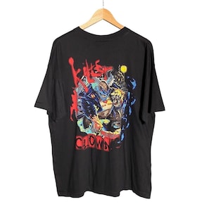 Op de afbeelding: Een zwart t-shirt met korte mouwen en een grafische print op de voorzijde. Het ontwerp toont een gestileerde illustratie van een clownfiguur met rode, blauwe en gele accenten. De tekst "KILLER CLOWNS" is gedrukt in een vet, verweerd rood lettertype.