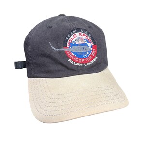 Puede incluir: Una gorra de béisbol gris carbón con visera beige. La parte frontal presenta un parche circular bordado con un helicóptero, una pequeña bandera estadounidense y el texto "TEAM U.S.A. POLO SPORT HELICOPTER SKI RALPH LAUREN" en letras blancas y rojas.